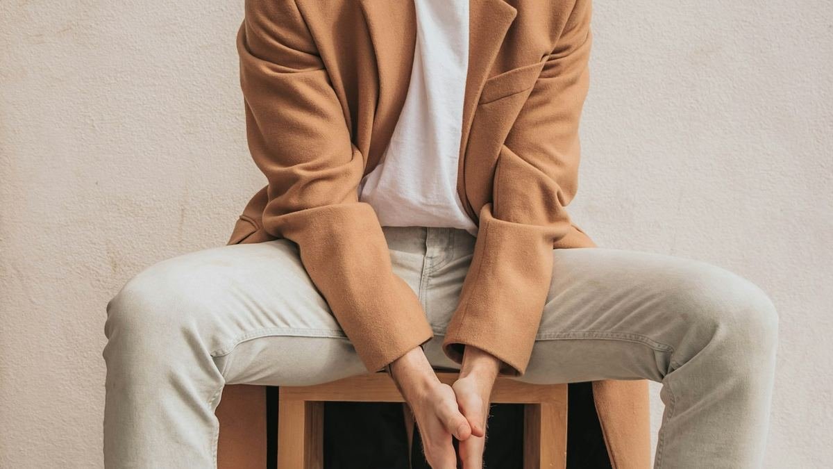 Person in a camel coat, white t-shirt and jeans seated against a plain wall